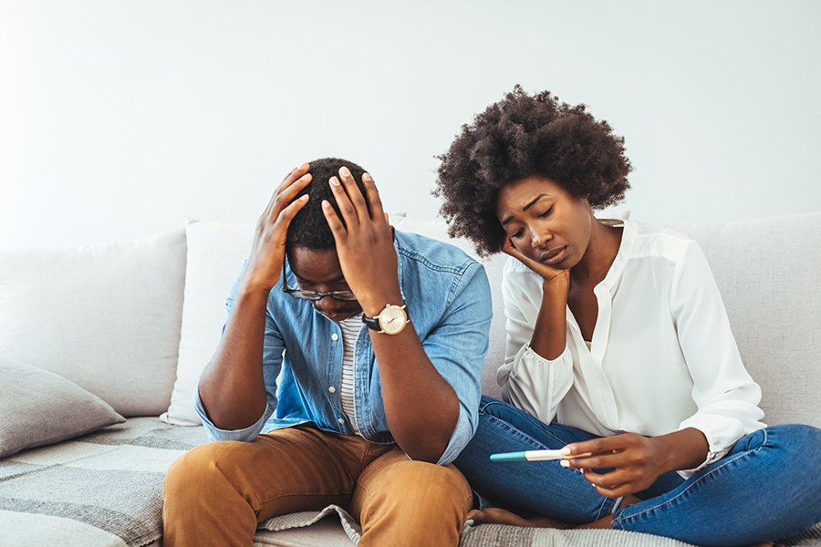 Upset man and woman sitting on couch apart, holding pregnancy te